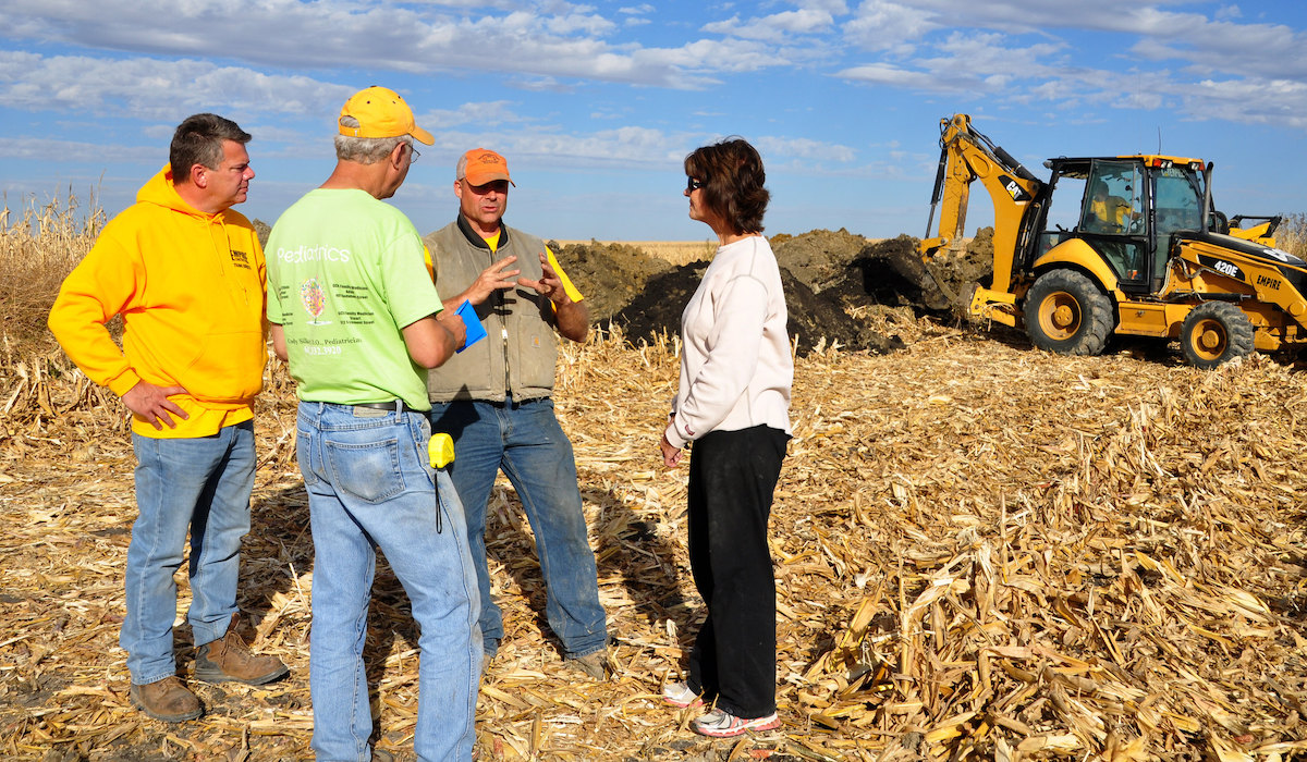 People standing and talking in farm field with bulldozer in background